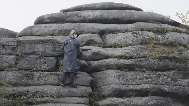 Rear view of a man in a long jacket and colored hair trying to climb a stony mountain. Male hiker climbing giant bouldrers on cloudy sky background.