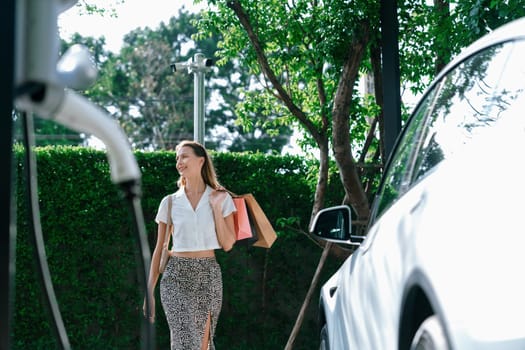 Young woman travel with EV electric car charging in green sustainable city outdoor garden in summer. Urban sustainability lifestyle by green clean rechargeable energy of electric BEV vehicle innards