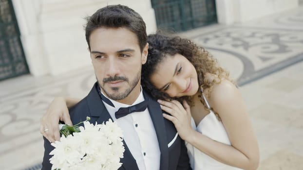 Newlyweds, caucasian bride and groom standing outdoors in the city street. Action. Woman in white dress with white bouquet lying on the shoulder of her man in suit