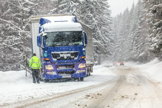 Jaraba, Slovakia - January 08, 2019: Unknown driver in green high visibility jacket standing next to truck, stopped next to slippery snow covered road, unable to move further, during heavy blizzard. 