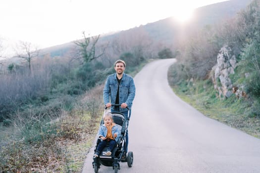 Smiling dad with a little girl in a stroller walks along the road on the mountainside. High quality photo