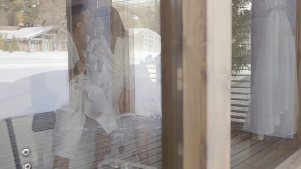 Young couple in love in bed with glasses full of champagne. View through the hotel room window, romantic evening of man and woman in bathrobes kissing each other