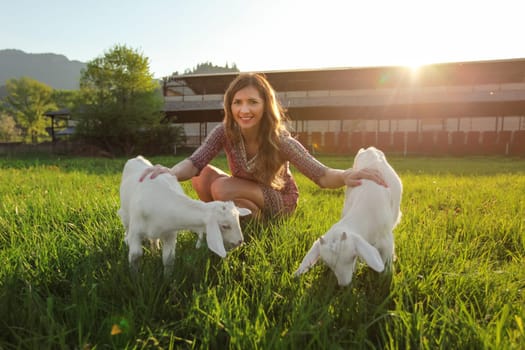 Young woman with two goat kids on spring meadow, wide angle photo, strong sun backlight over farm in background