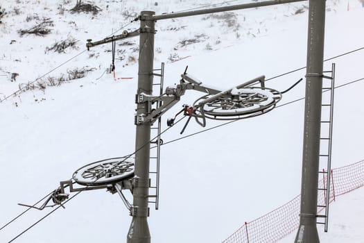 Large metal wheels for steel cables on top of ski lift supporting pillar, snow covered piste in background