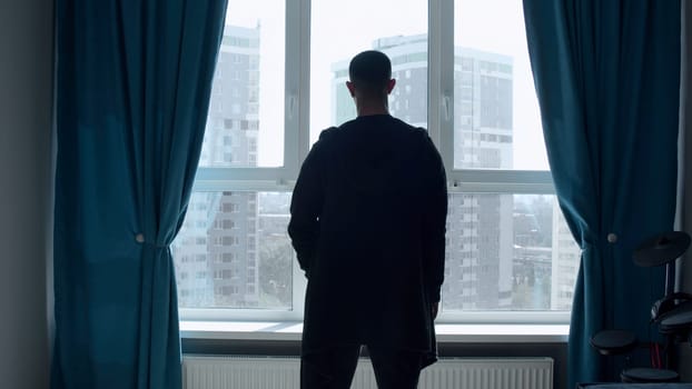 An adult man standing in the room. Media. A beautiful slender silhouette of a man who stands in a bright room with a large window and looks into the distance. High quality 4k footage