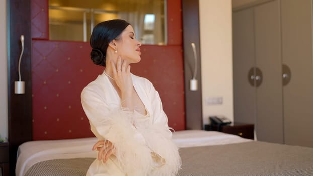 Portrait of a young brunette woman sitting on the bed in a hotel room or bedroom. Action. Beautiful lady indoors in white robe with feathers