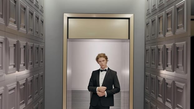 Handsome stylish groom in the hotel interior posing in front of the camera. Action. Man in black suit and white shirt standing by the wall in the end of the corridor