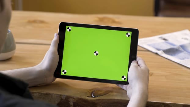 Young guy holding the tablet with touching green screen with tracking marks, motionless. Modern device or ipad with big chromakey monitor on wooden table background.