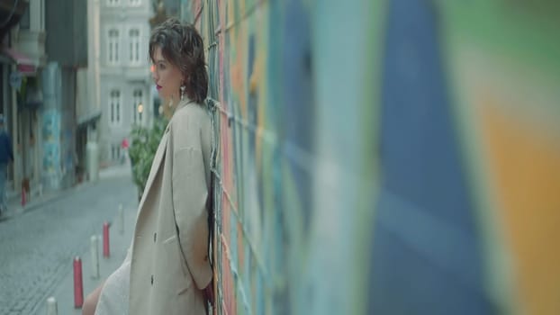 Stylish young woman stands at street wall. Action. Side view of attractive young woman against wall. Woman is standing on street by wall.