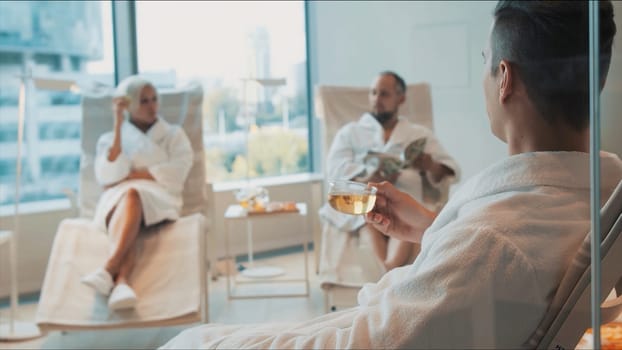 People relaxing at the spa salon. Video. Two men and a woman in white bathrobes having a rest with tea, newspaper inside a spacious room with panoramic windows