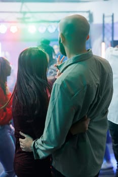 Young couple hugging and standing on dancefloor while partying in nightclub. Boyfriend holding girlfriend waist while clubbing together, having fun and enjoying nightlife at discotheque