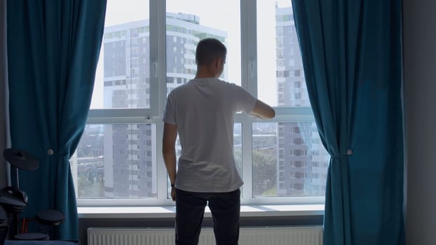 Young man standing near the window in a room with blue curtains. Media. Man waiting for something, looking at watch