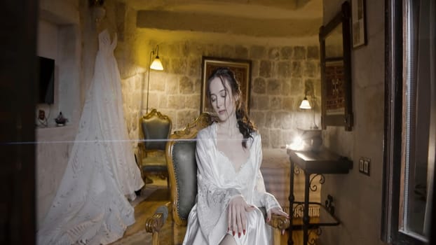 Morning of a bride. Action. Beautiful bride in a hotel room in a white silk nightdress with a wedding dress behind her