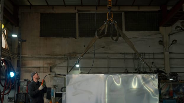 Manipulator with metal cables and operator transporting a massive heavy metal rectangular detail at steel plant. Creative. Modern equipment at industrial workshop factory