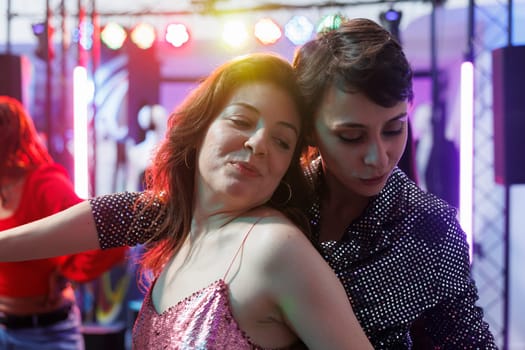 Smiling women hugging while dancing together at nightclub party. Young romantic girlfriends couple relaxing and moving on crowded dancefloor while clubbing at discotheque