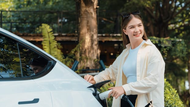 Young woman recharge her EV electric vehicle at green city park parking lot. Urban sustainability lifestyle for environmental friendly EV car with battery charging station. Expedient