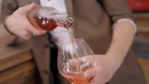 Close up of a man in beige suit pouring rose wine into a transparent glass. Action. Male sommelier pouring fruit wine at a bar