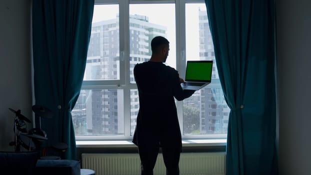 Rear view of a man standing by the window with a laptop in his hands. Media. Computer with green chroma key screen