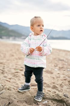 Little girl stands on the beach with a stick in her hands and looks into the distance. High quality photo