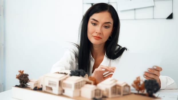 Closeup of young beautiful architect compares blueprints with architectural model with house model and architectural equipment placed on table at meeting room. Creative design concept. Immaculate.