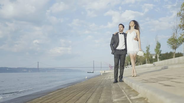 Beautiful couple in love hugs on embankment. Action. Elegant couple in suits during date on embankment. Beautiful couple of lovers tenderly embrace on embankment on sunny day.