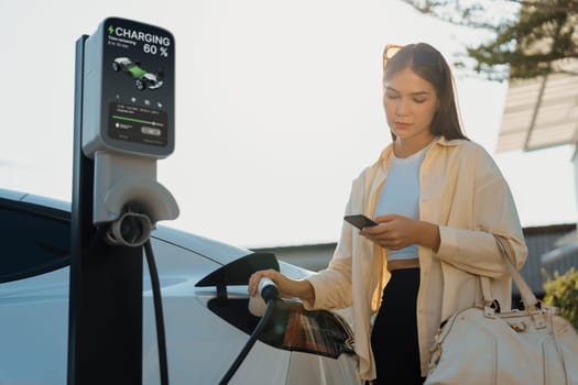 Young woman holding shopping bag and use smartphone to pay for electricity for recharging EV car battery from charging station at city mall parking lot. Modern woman go shopping by eco car. Expedient