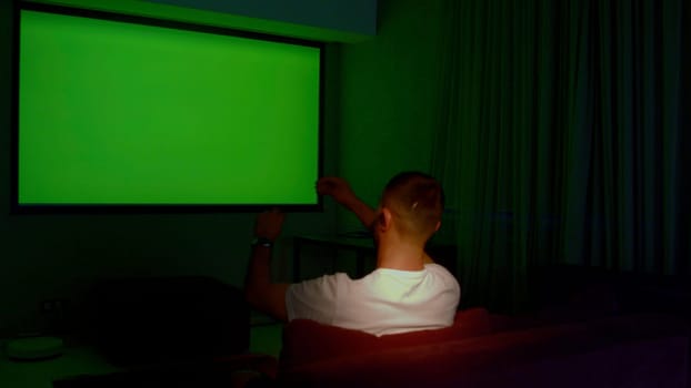Man watching TV with green screen in living room, back view. Media. Man switching channels with hand gestures, gesture interface, modern technology