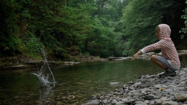 Boy child squatting in front of a river and throwing stones into the water. Creative. Kid exploring nature on a summer day