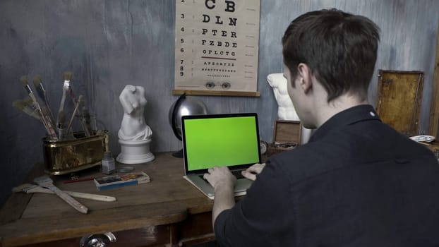 Back view of an artist in old fashioned art studio sitting at the wooden table and working on his laptop with chromakey green screen. Man in the stylized room.
