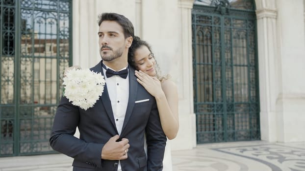 Newlyweds, caucasian bride and groom standing outdoors in the city street. Action. Woman in white dress with white bouquet lying on the shoulder of her man in suit