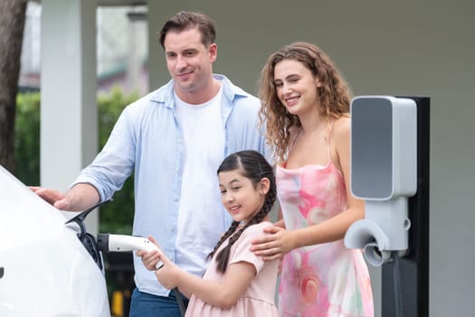 Happy little young girl learn about eco-friendly and energy sustainability as she help her family recharge electric vehicle from home EV charging station. EV car and modern family concept. Synchronos