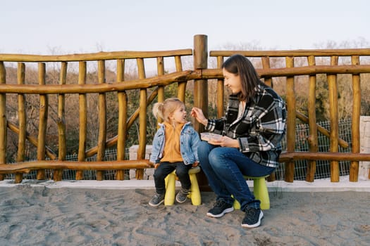 Mom feeds a little girl from a spoon while sitting on chairs near a wooden fence. High quality photo