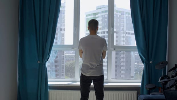 Young confident man standing looking through the window at home with his arms folded. Media. Rear view of a young male model