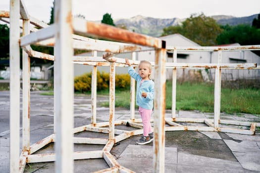 Little girl stands near a metal labyrinth and holds on to a beam with her hand. High quality photo