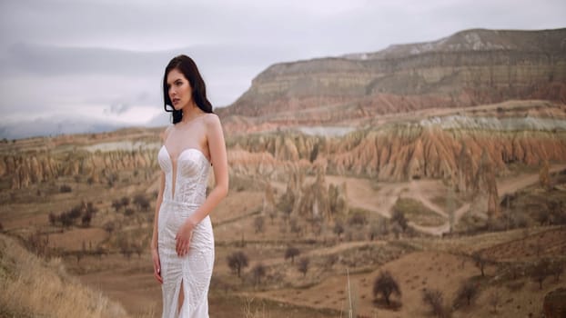 Woman wearing bride dress stands on the canyon landscape background. Action. Beautiful young brunette woman in front of picturesque valley
