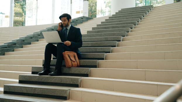 Skilled investor working or planing strategy by using laptop at stair. Professional business man wearing suit while working and typing data analysis by using laptop and talking to phone. Exultant.