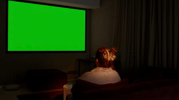 Woman watching television in living room late in the evening. Media. Young girl sitting on sofa against tv with chroma key green screen, back view