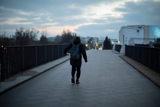 Guy walks across bridge in evening. Man in town at night. Man with backpack in black clothes. Suspicious person.