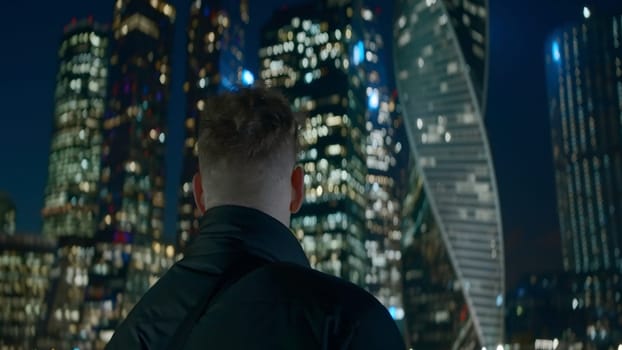 Man looks at modern glowing skyscrapers at night. Action. Young man on background of bright glowing skyscrapers at night. Man looks at skyscrapers glowing at night in city.
