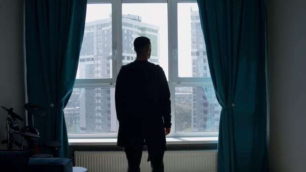 Rear view of a dressed man standing indoors in a living room by the window. Media. Silhouette of a man looking through the window