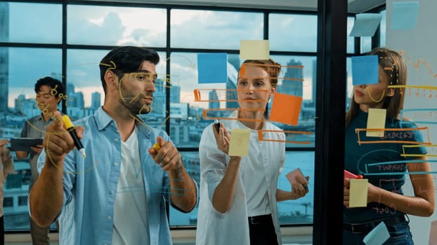 Professional business team brainstorm, sharing, thinking idea while skilled male leader write on sticky notes at glass wall. Skilled manager placed business plan on glass wall at office. Tracery