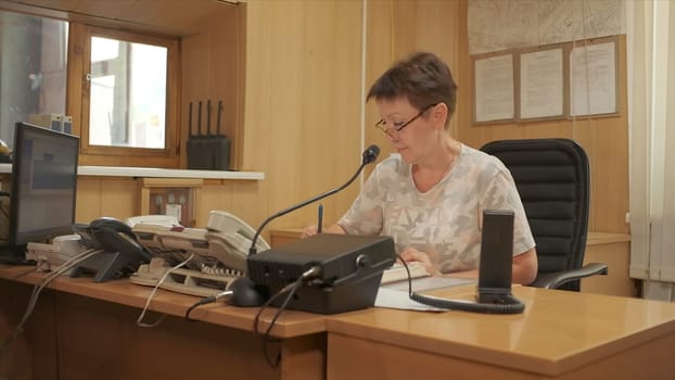 Woman is sitting in office and answering phone. Clip. Elderly woman answers phone. Office with woman for phone calls for help.