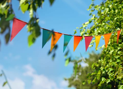 colorful pennant string decoration in green tree foliage on blue sky, summer party background.