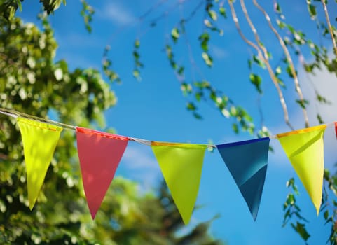 colorful pennant string decoration in green tree foliage on blue sky, summer party background.