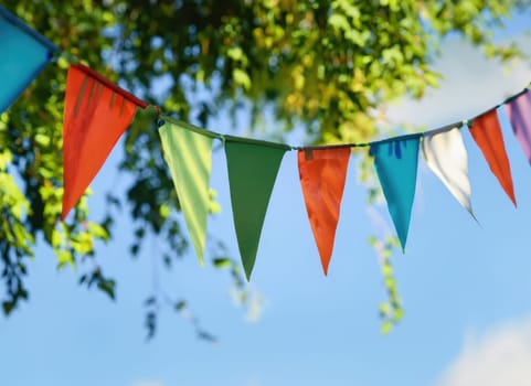 colorful pennant string decoration in green tree foliage on blue sky, summer party background.