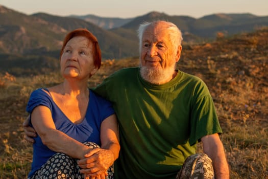 An elderly couple sits on a mountain with their backs with a beautiful view of the mountains in the distance
