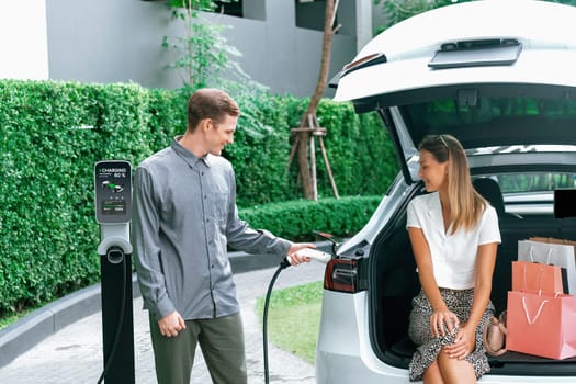 Young couple travel with EV electric car charging in green sustainable city outdoor garden in summer shows urban sustainability lifestyle by green clean rechargeable energy of electric vehicle innards