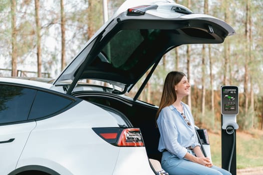 Young woman recharging battery for electric car during autumnal road trip travel EV car in autumnal forest. Eco friendly travel on vacation during autumn with electric vehicle. Exalt