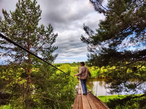 A girl on an old wooden suspension bridge among spruce or pine trees in nature. A tourist on a trip and landscape with trees on a cloudy autumn or spring day