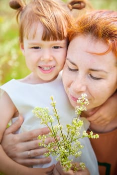 Happy female family with mother and daughter on green and yellow meadow full of grass and flower. Woman with red hair and blonde girl having fun, joy and hug in sunny summer day. Concept family love